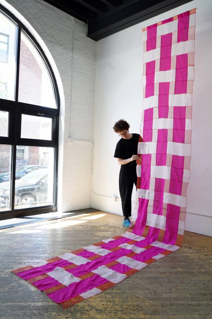 Amy stands next to a large pink and white transluscent textile art work that hangs down the white studio wall and across the wooden floor