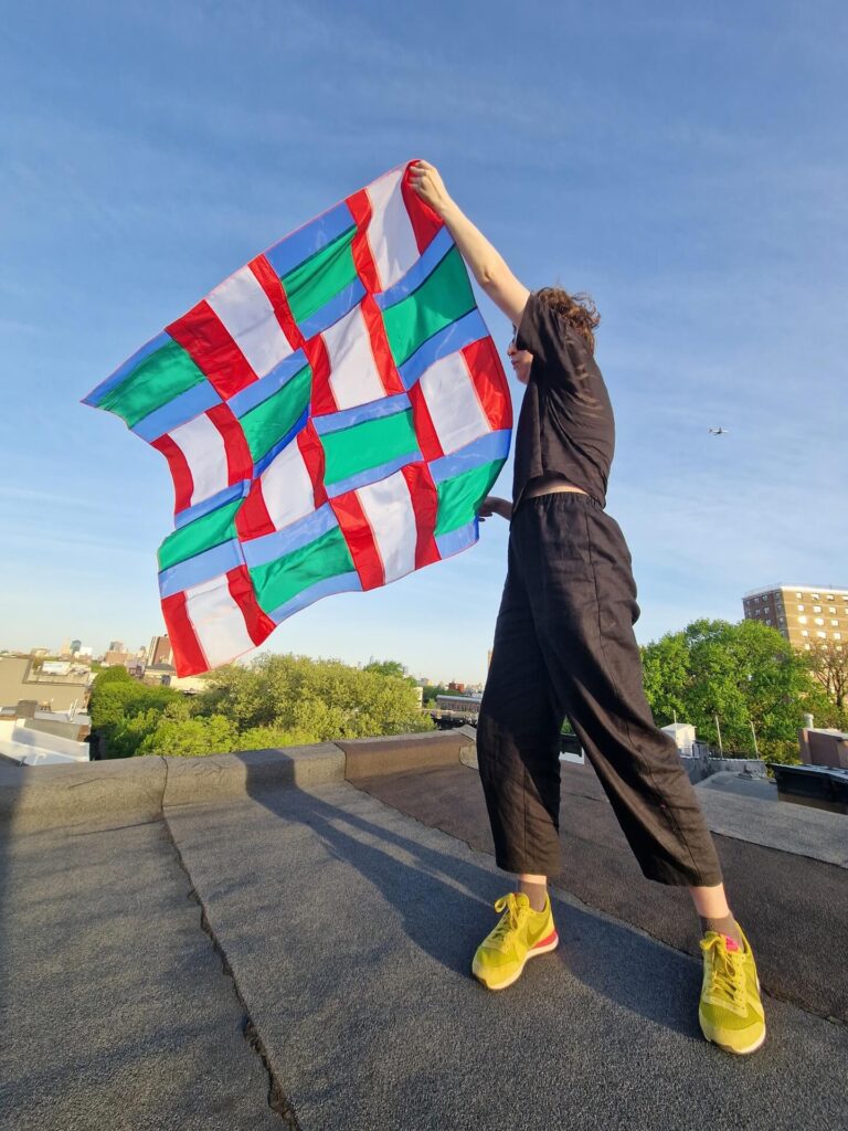 Amy standing on a Brooklyn rooftop holding a piece of multicoloured textile artwork in the breeze